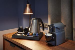 Hospitality trays for hotels in hotel room with Metallic KEttle and black mugs on sleek black tray. Coffee pod machine on counter, with cup being used to dispense a coffee pod coffee